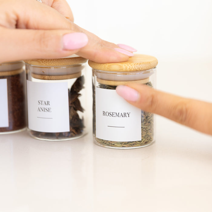 Three small glass jars with wooden lids labeled 'Star Anise', 'Cinnamon', and 'Rosemary' on a white background.