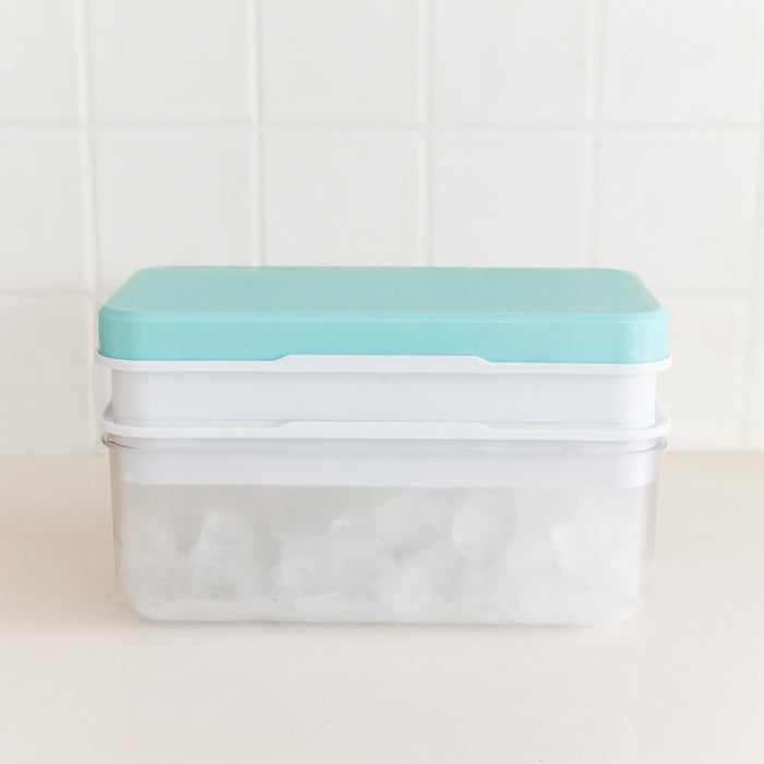 A clear plastic ice cube tray container with a light blue lid and white inner tray, filled with ice cubes, set against a white tiled background.