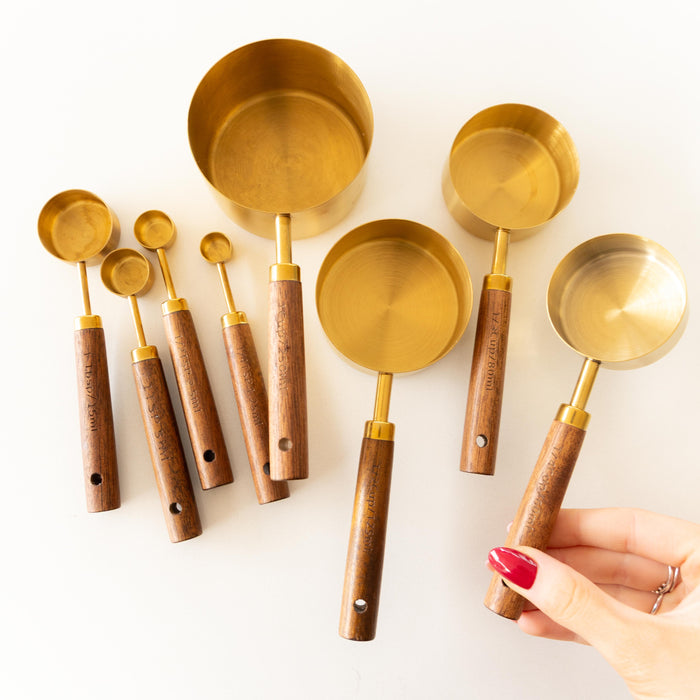 A set of brass measuring cups and spoons with wooden handles arranged on a white background, with a hand holding one of the larger cups.