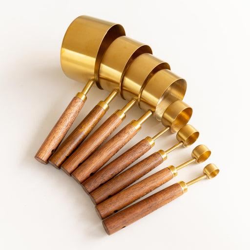 A set of nested measuring cups with shiny gold brass bodies and light brown wooden handles, arranged diagonally on a clean white background.